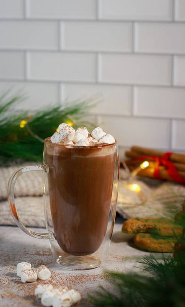 hot chocolate in a glass mug with mini marshmallows on grey table with cookies, mandarins and cinnamon sticks, blurred xmas decoration background . High quality photo - Photo, Image