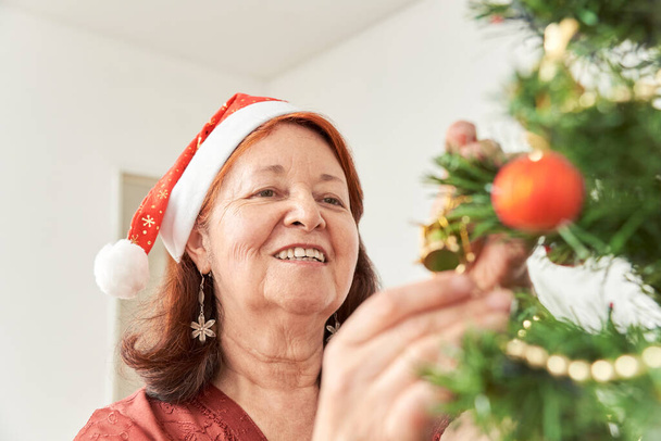 Gelukkige oudere Spaanse vrouw glimlachend terwijl ze een kerstboom versiert met een rode kerstmuts. De vreugde van de vakantie. - Foto, afbeelding