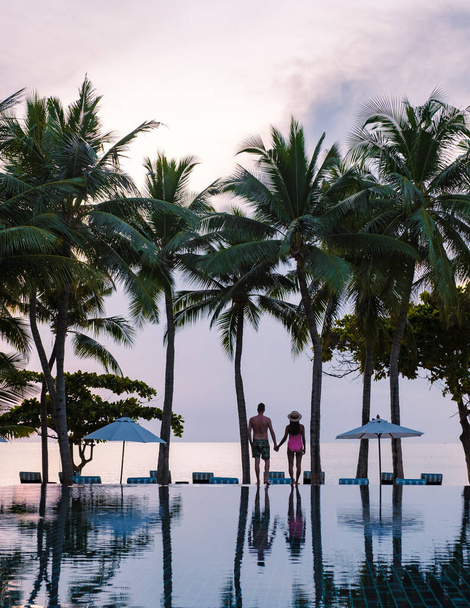 Un par de hombres y mujeres relajándose junto a la piscina en camas de playa sillas, piscina tropical y piscina con palmeras en la playa con vistas al océano - Foto, Imagen