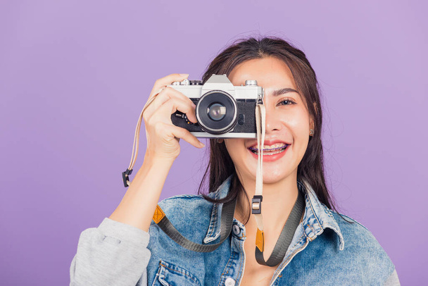 Portrait happy beautiful young woman smile excited wear denims photographer taking a picture and looking viewfinder on vintage photo camera ready to shoot, studio shot isolated on purple background - 写真・画像