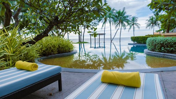 basen tropikalny z palmami i leżakami plażowymi w luksusowym kurorcie, basen z palmami na plaży z widokiem na ocean. krzesła plażowe z ręcznikiem plażowym przy basenie - Zdjęcie, obraz