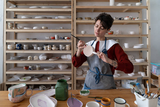 工房絵画マグカップの作業台に立つ若いクリエイティブ・クラフツマンシップ。陶芸のワークショップで創造性を表現し、女性陶芸家が販売するためのユニークなセラミック作品を作成します。中小企業 - 写真・画像