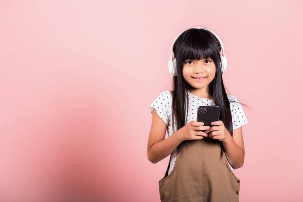 Asian kid 10 years enjoying listening music from mobile phone wear wireless headset at studio shot isolated on pink background, Happy child girl lifestyle use smartphone listen music with headphones - Fotó, kép