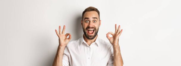 Close-up of amazed caucasian guy agreeing, showing okay signs and looking impressed, recommending product, white background. - Фото, изображение