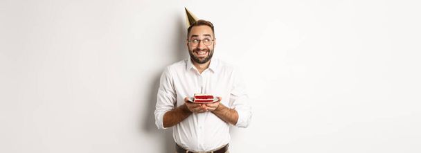 Svátky a oslavy. Happy man having birthday party, making wish on b-day cake and smiling, standing against white background. - Fotografie, Obrázek