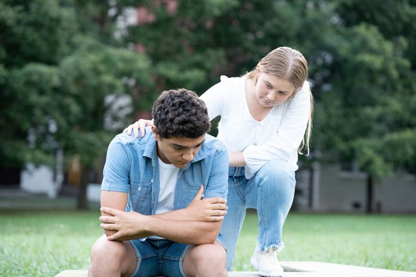 Ένα έφηβο κορίτσι παρηγορεί ένα θλιμμένο ισπανόφωνο αγόρι σε ένα πάρκο. Έννοια πένθους, κατάθλιψης και άγχους. - Φωτογραφία, εικόνα