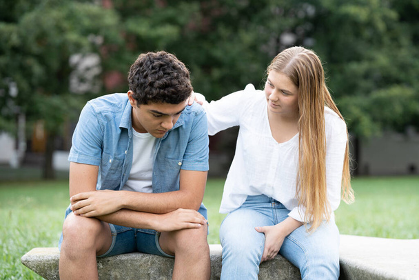 Teenager girl comforting sad boyfriend sitting on a bench outdoors in a park. Mourning and depression assistance concept - Photo, Image