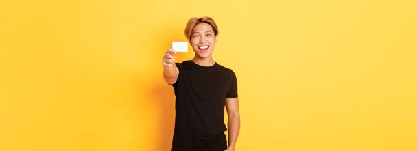 Веселий красивий азіатський хлопець посміхається задоволений і показує кредитну картку, стоячи жовтим фоном
. - Фото, зображення