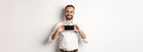 眼鏡をかけたハンサムな髭の男、携帯電話のアプリケーションを実証し、スマートフォンの画面を示し、白い背景の上に立って. - 写真・画像