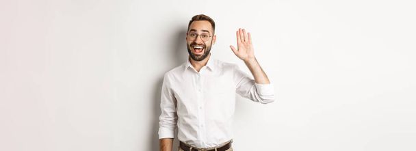 Friendly smiling man in glasses saying hello, waving hand in greeting, standing over white background. - Photo, Image