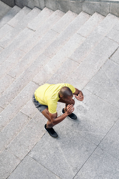 Aerial view of an African American sportsman with yellow t-shirt and shorts doing squats on stairs outdoors. - Φωτογραφία, εικόνα