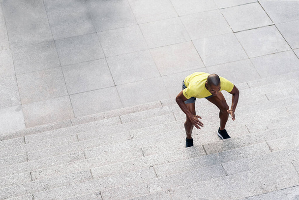 Bovenaanzicht van een Afro-Amerikaanse sportman met geel t-shirt en korte broek die buiten op de trap hurkt. - Foto, afbeelding