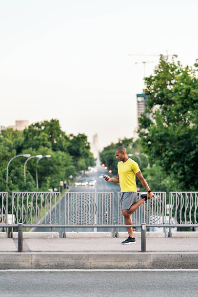 African American man stretching his left leg and holding phone in the footbridge at day. - Foto, Bild