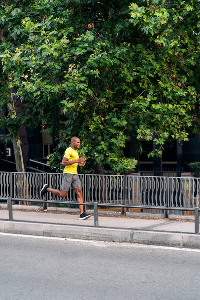 African American sportsman with yellow t-shirt and shorts sprinting on sidewalk in the city outdoors with trees and green leaves behind. - Foto, afbeelding