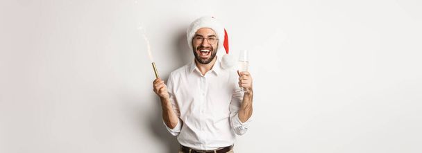 Wintervakantie en feest. Knappe man met baard met nieuwjaarsfeest, met vuurwerk en champagne, met kerstmuts op, witte achtergrond. - Foto, afbeelding