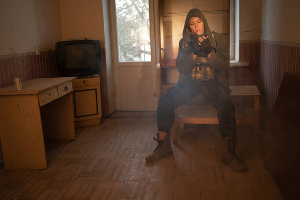 Περιβαλλοντική καταστροφή. Μετα-αποκαλυπτική γυναίκα επιζών καπνίζει σε εγκαταλελειμμένο κτίριο. Ένας μοναχικός ήρωας που φοράει στρατιωτική στολή καθισμένος στον αντίχειρα. - Φωτογραφία, εικόνα