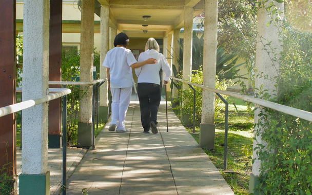 病院の庭にウォーカーの棒で高齢者の女性を助けるアジアの医師。リハビリテーションの概念. - 写真・画像