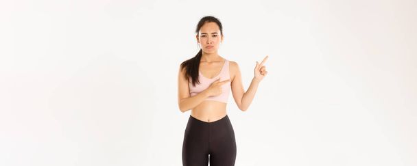 Sport, wellbeing and active lifestyle concept. Complaining sad asian girl in activewear, sulking and frowning upset as pointing upper left corner, end of sale offer, standing white background. - Foto, imagen
