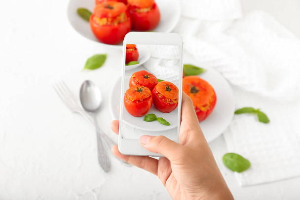 Fotógrafa de comida con teléfono móvil tomando fotos de sabrosos tomates rellenos en mesa blanca - Foto, imagen