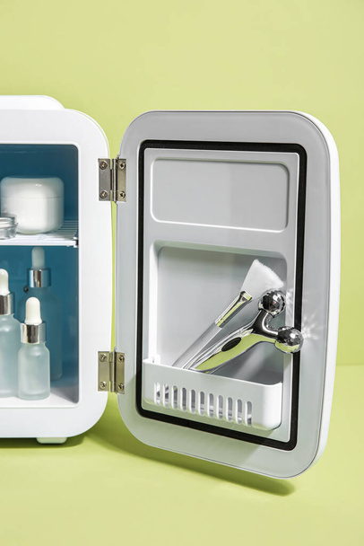 Невеликий холодильник з натуральною косметикою, пензлем та масажним інструментом на зеленому фоні
 - Фото, зображення