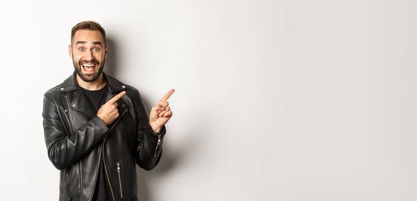Excited, handsome man in black leather jacket, pointing fingers right at promo offer, showing logo, white background. - Fotoğraf, Görsel