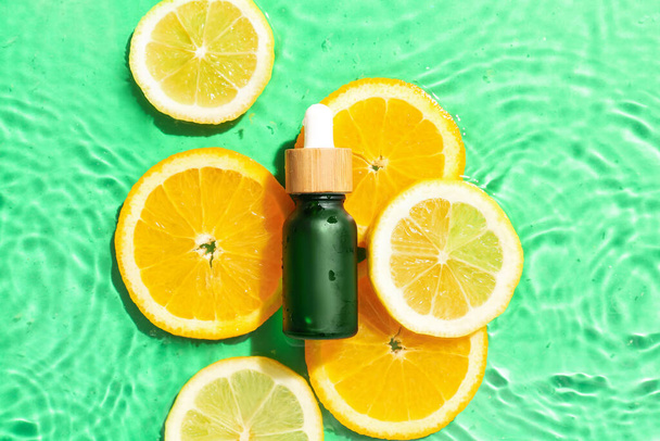 Bottle with vitamin C serum and slices of citrus fruits in water on color background - Fotó, kép