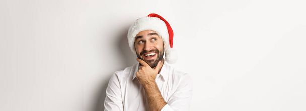 Impreza, ferie zimowe i koncepcja świętowania. Zbliżenie szczęśliwy człowiek planowania listy prezentów na Boże Narodzenie, noszenie czapki Mikołaja, patrząc w lewym górnym rogu przemyślane, białe tło. - Zdjęcie, obraz