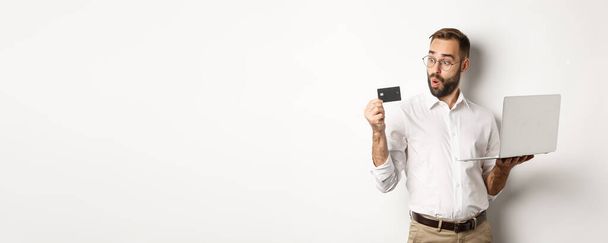 Compras online. Empresário surpreendente segurando laptop, olhando impressionado com cartão de crédito, de pé sobre fundo branco. - Foto, Imagem