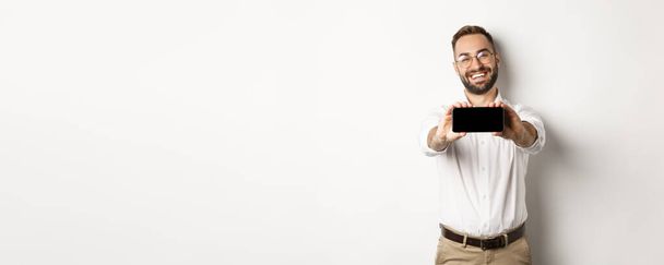 Felice uomo d'affari che mostra lo schermo mobile, tenendo il telefono orizzontalmente, in piedi soddisfatto sullo sfondo bianco. - Foto, immagini