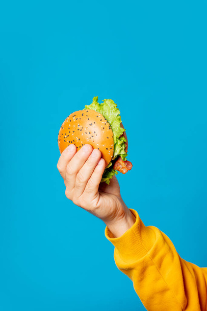 Male hand in yellow clothes hold hamburger on blue background - Foto, Imagen