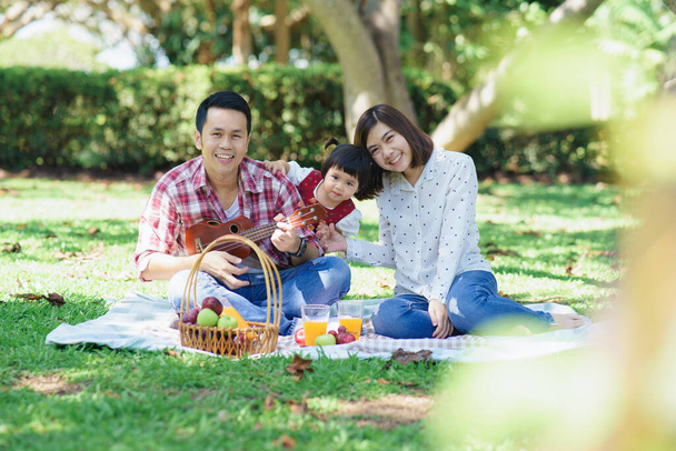 Family with children enjoying picnic in spring garden. Parents and kids having fun eating lunch outdoors in summer park. Mother and father and daughter playing ukulele in garden - Photo, Image