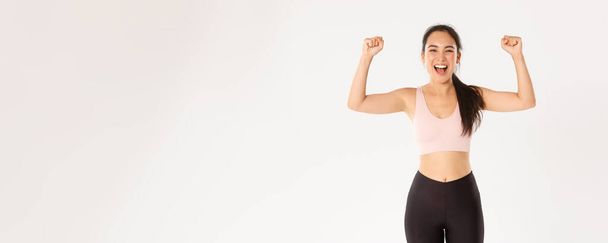 Sport, wellbeing and active lifestyle concept. Empowered and strong asian female athlete, fitness girl encourage herself for good workout, fist pump and shouting upbeat, white background. - Photo, Image