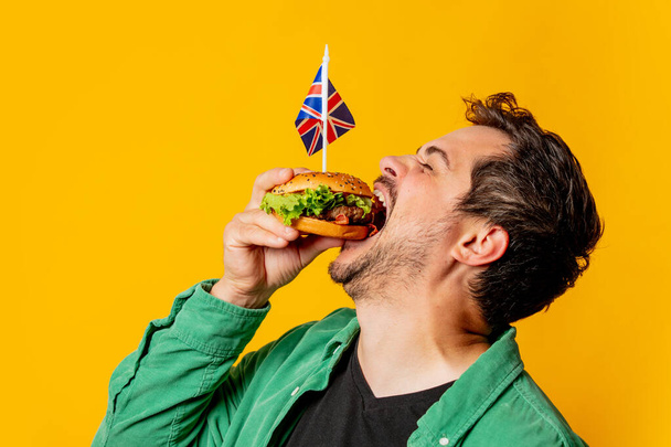 Улыбающийся мужчина с флагом Великобритании и бургером на желтом фоне - Фото, изображение
