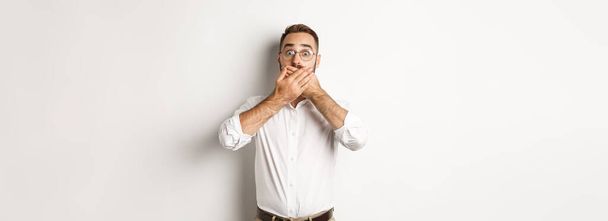 Shocked man gasping and looking at something in awe, covering mouth with hands, white background. - Foto, afbeelding