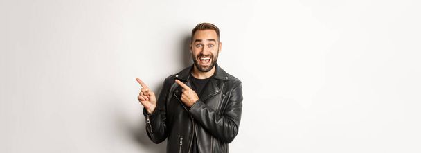 Excited, handsome man in black leather jacket, pointing fingers right at promo offer, showing logo, white background. - Photo, Image