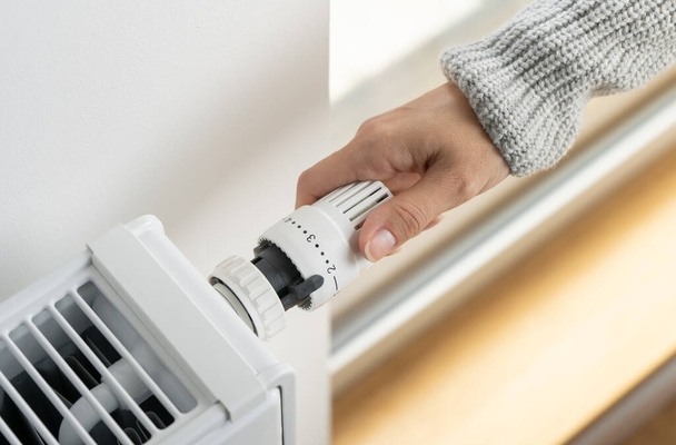 Una mujer en un suéter caliente gira el regulador de la válvula del radiador termostático a la posición mínima para ajustar el modo de calefacción económica de la habitación. Calefacción del apartamento en la estación fría. - Foto, imagen