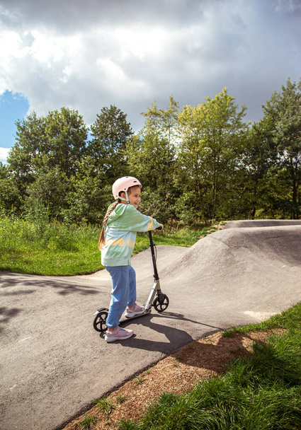  Портрет милой маленькой кавказской школьницы носить шлем наслаждаться весело кататься на скутере на асфальтированной дорожке в уличном парке на открытом воздухе в солнечный день. Занятия спортом для детей вне игры - Фото, изображение