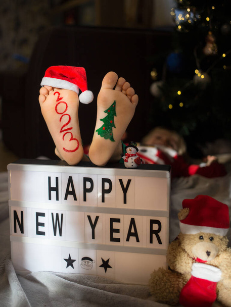 Noel hazırlığı konsepti. pozitif, şımartıcı, esprili. 2023 numara, çocuğun çıplak ayaklarıyla yazılmıştır. Yeni yılınız kutlu olsun. Yeni yıl teması üzerine neşeli samimi resimler - Fotoğraf, Görsel