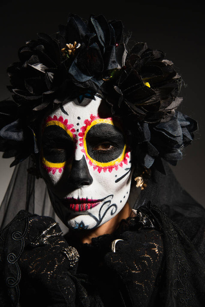Portrait of woman in wreath and catrina makeup looking at camera on black background  - Φωτογραφία, εικόνα