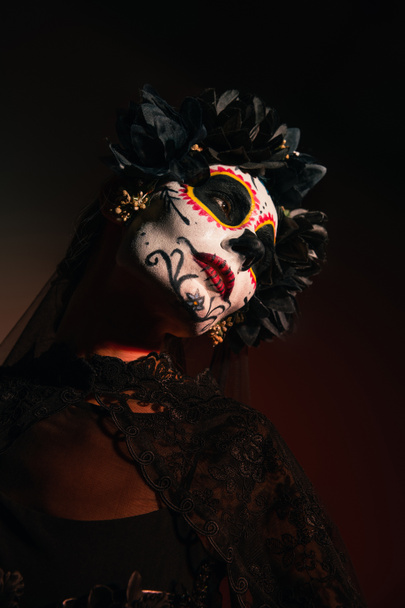Mujer en guirnalda con flores negras y maquillaje catrina mirando hacia otro lado sobre fondo borgoña  - Foto, imagen