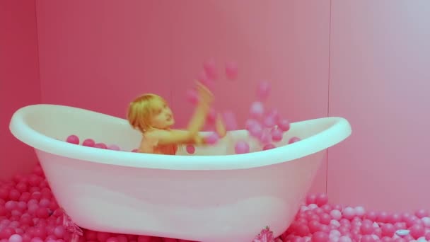 Happy Little hermosa rubia caucásica chica se sienta junto al baño y lanza bolas de color rosa. Feliz infancia. primer plano, enfoque suave - Metraje, vídeo