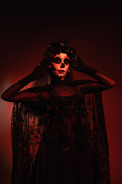 Mujer en traje negro y maquillaje de santa muerte tocando corona negra sobre fondo borgoña con iluminación roja - Foto, Imagen