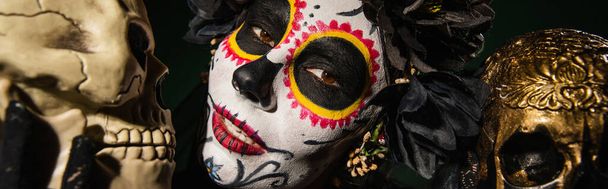 Portrait of woman in santa muerte makeup and black wreath looking at camera near skulls isolated on black, banner  - Photo, Image