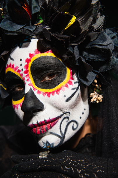 Portrait of woman with sugar skull makeup and dark wreath looking at camera isolated on black  - Photo, Image