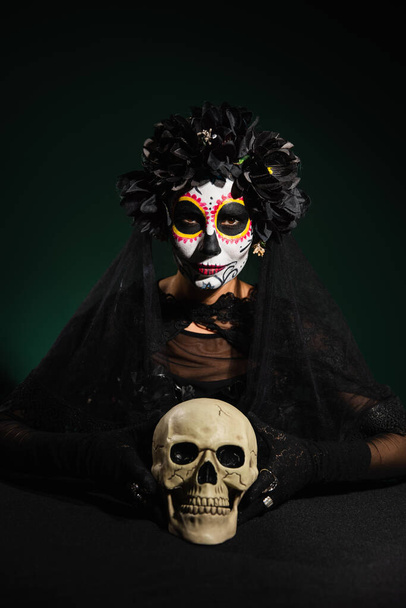 Mulher em grinalda com véu e maquiagem catrina olhando para a câmera perto do crânio no fundo verde escuro  - Foto, Imagem
