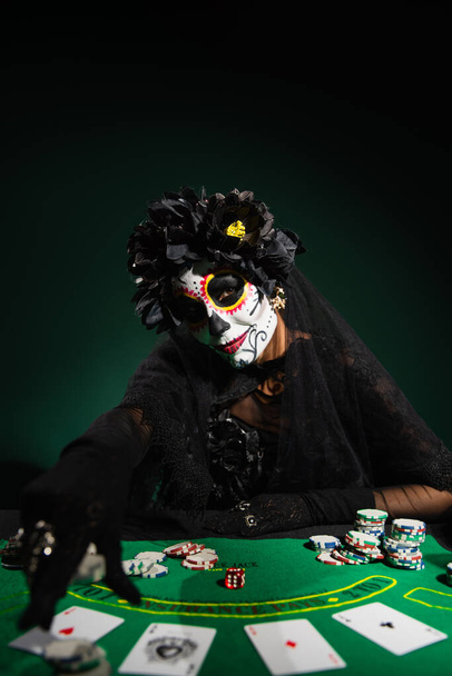 KYIV, UKRAINE - SEPTEMBER 12, 2022: Woman in mexican day of dead costume near playing cards on dark green  - Fotografie, Obrázek