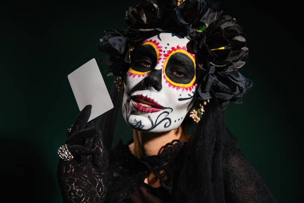Portrait of woman in santa muerte costume and makeup holding card isolated on dark green  - Photo, Image