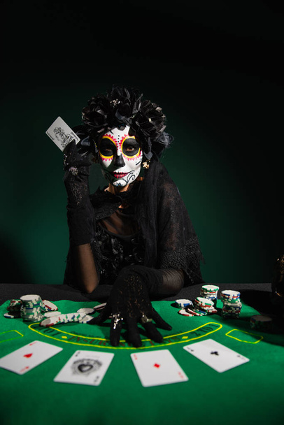 KYIV, UKRAINE - SEPTEMBER 12, 2022: Woman with sugar skull makeup and costume holding playing card on dark green - Fotoğraf, Görsel