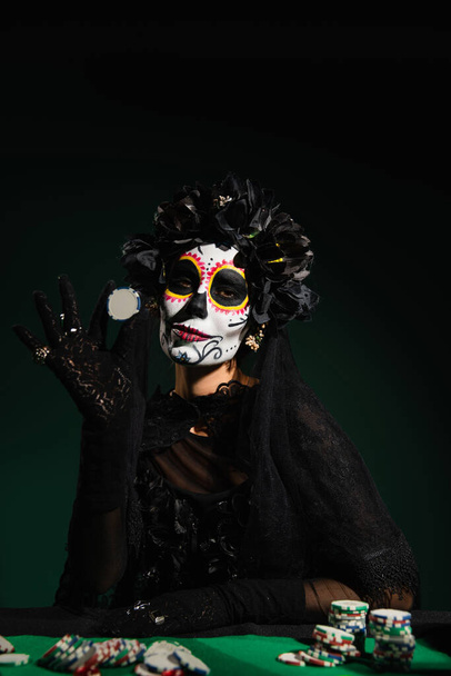 Woman in santa muerte costume and wreath holding playing chip on dark green background  - Photo, Image