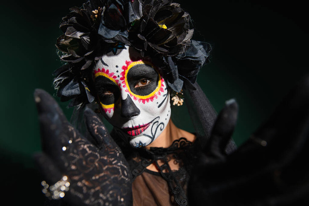 Woman in sugar skull makeup and wrench with black flowers looking at camera on dark green background  - Photo, Image